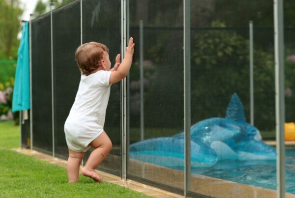 A baby leaning on a fence surrounding a backyard pool.