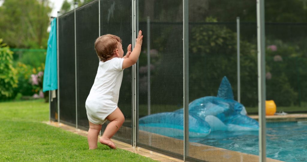 A baby leaning on a fence surrounding a backyard pool.